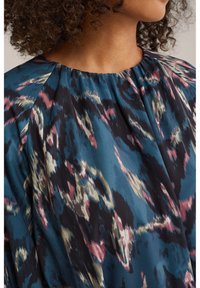 Top din țesătură imprimată cu un fundal teal și modele abstracte în nuanțe de roz, crem și negru. Are un guler rotund și o textură moale.