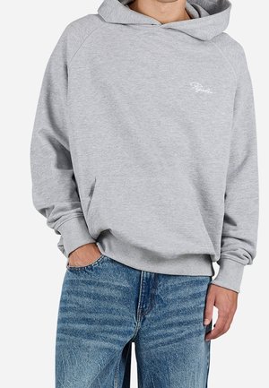 Person iført en lysegrå hættetrøje med lomme foran og lille hvid tekstlogo, kombineret med blå jeans, står med den ene hånd i lommen.