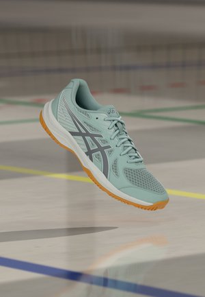 Ανοιχτό μπλε αθλητικό παπούτσι Asics με πορτοκαλί σόλα να αιωρείται πάνω από το πάτωμα γυμναστηρίου με πολύχρωμες γραμμές στο γήπεδο.