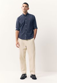 Marineblauw overhemd met knopen en opgerolde mouwen, beige broek en zwarte veterschoenen. Voorzien van een geborduurd rood logo op de borst van het overhemd.