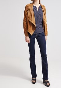 Hnědá semišová asymetrická bunda přes námořnické puntíkované tričko, spárovaná s tmavými džíny a černými platformovými sandály. Hladké textury a vrstvený design.