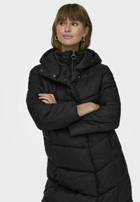 Parka noire rembourrée avec col haut, fermeture éclair à l'avant et capuche. Présente un motif matelassé avec des coutures diagonales et des boutons-pression.