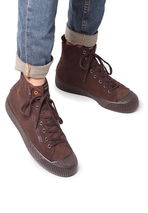 Zapatillas altas de ante marrón con suela de goma texturizada, que cuentan con ojales de metal y cordones. Se pueden ver unos jeans de mezclilla ajustados.