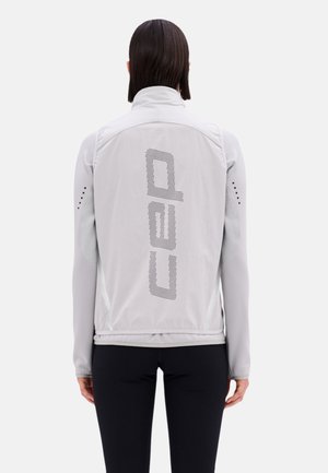 Persona che indossa una giacca grigio chiaro a maniche lunghe con testo nero traforato "cap" sulla schiena, abbinata a pantaloni neri, vista di spalle.