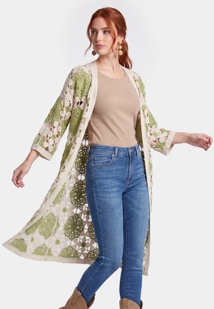 Kvinde med rødt hår iført beige top, blå jeans, beige og grøn hæklet cardigan og brune støvler, poserer med armene let strakt ud.