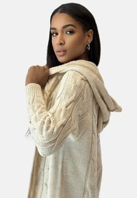 Krämfärgad kabelstickad kofta med huva, med texturerade mönster och långa ärmar, hängande över modellens axlar.