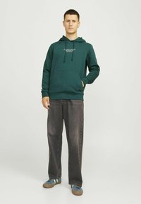 Grüne Kapuzenjacke mit Kordelzug, Fronttasche und Logo. Getragen mit lockeren, dunkelgrauen Hosen und blauen Sneakers mit weißen Streifen.