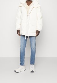 Bílá péřová bunda s velkými kapsami, kapucí a elastickými manžetami, spárovaná se světle modrými džíny a bílými teniskami s šedými akcenty.