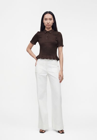 Femme debout, une main sur la hanche, portant un haut marron texturé à manches courtes et un pantalon blanc large taille haute sur fond blanc.