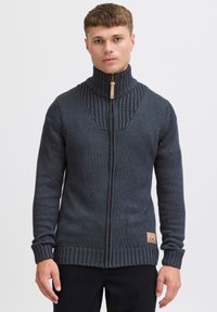 Mörkgrå zip-up stickad tröja med hög krage, ribbat mönster, kontrasterande paneldesign och märkespatch längst ner till vänster.