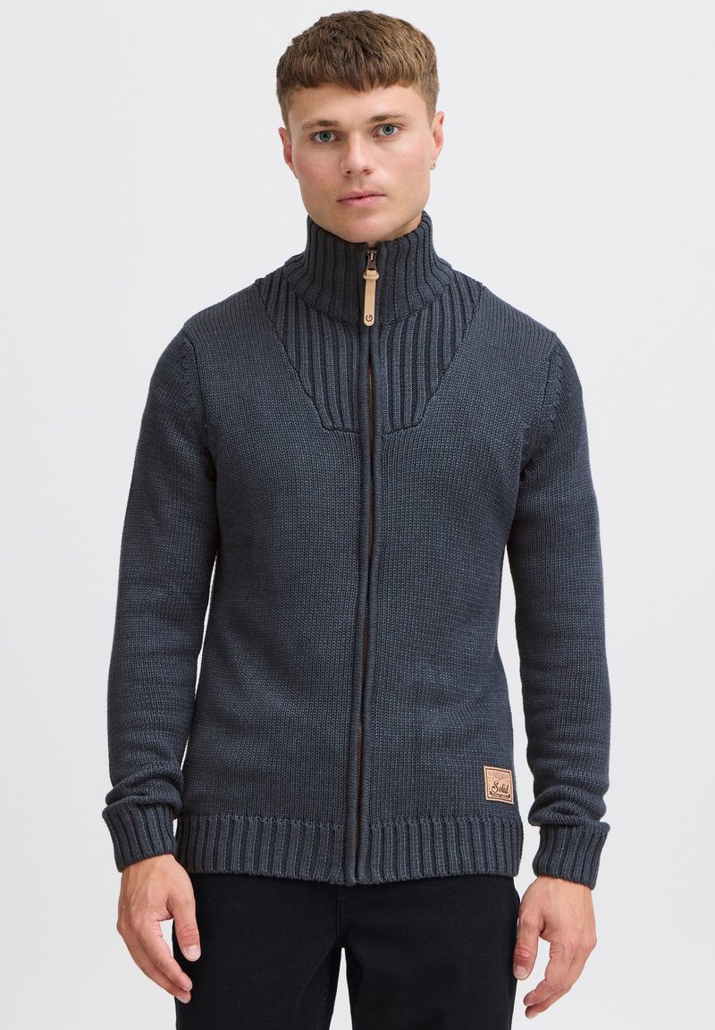 Mörkgrå zip-up stickad tröja med hög krage, ribbat mönster, kontrasterande paneldesign och märkespatch längst ner till vänster.