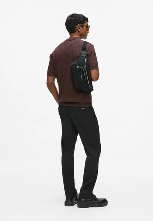 Man draagt een bruin shirt met korte mouwen, zwarte broek, zwarte schoenen, zonnebril, draagt een zwarte crossbodytas over zijn schouder, kijkt zijwaarts.