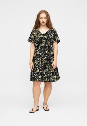 Kobieta o długich czerwonych włosach, ubrana w czarną kwiecistą sukienkę do kolan i czarne sandały, stoi na tle jednolitego jasnego tła.