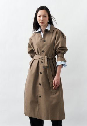 Žena v zapnutém, opásaném světle hnědém trenčkotu přes světle modrou košili s límečkem, stojící před jednobarevným bílým pozadím.