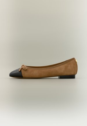 Scarpa da balletto piatta con corpo in camoscio beige, punta nera appuntita, piccolo fiocco beige e tacco basso nero su sfondo neutro.