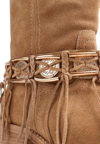 Botte en daim beige avec une lanière en cuir tressé décorative, des conchos floraux en métal, des barrettes dorées et des pompons suspendus près de la cheville.