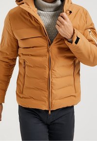 Jasnobrązowa puchowa kurtka z kapturem, zapinana na zamek z przodu, z bocznymi kieszeniami i prążkowanym kołnierzem. Gładka struktura i pikowany wzór.