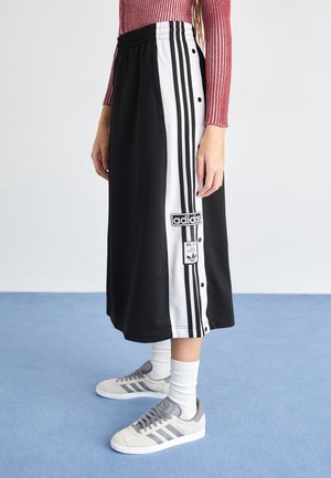 Frau trägt schwarzen und weißen Adidas-Midirock mit Logos und grauen Adidas-Sneakern auf blauem Boden, seitlich stehend.
