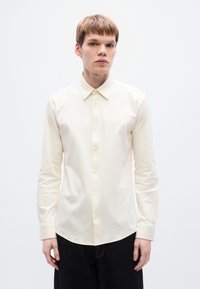 Μπλούζα μακριάς μανίκας σε κρεμ απ όχρωση, με μυτερό γιακά, κουμπιά μπροστά και διακριτική λεπτομέρεια λογότυπου στο στήθος· υφή υφάσματος λεία.