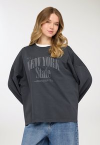 Felpa oversize grigio scuro con collo rotondo, caratterizzata dalla scritta "NEW YORK State" e maniche lunghe comode, abbinata a jeans denim azzurri.