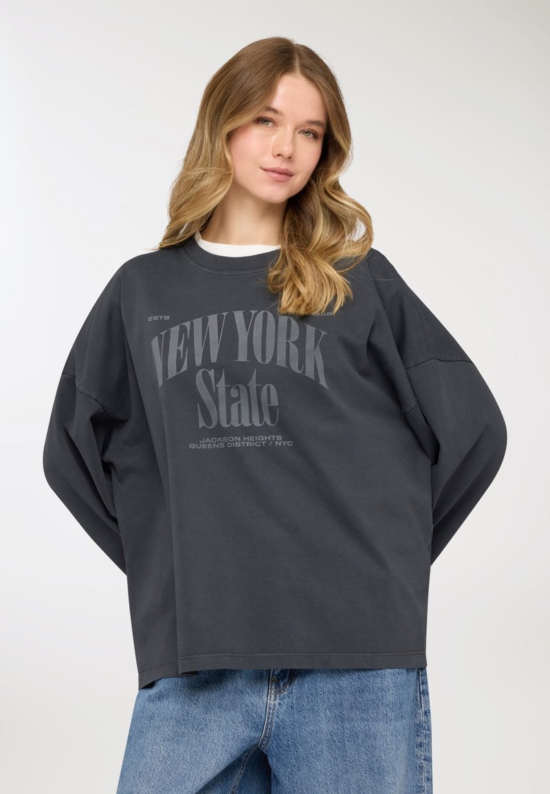 Felpa oversize grigio scuro con collo rotondo, caratterizzata dalla scritta "NEW YORK State" e maniche lunghe comode, abbinata a jeans denim azzurri.