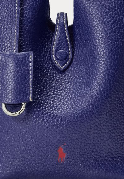Close-up of dark blue pebbled leather bag with stitched tab and silver ring detail, featuring a small red polo player logo.