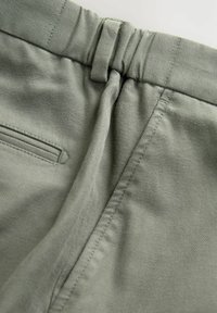 Material din bumbac de culoare verde deschis, cu o textură netedă, detalii de coasere, un brâu elastic și un buzunar lateral cu o finisare subtilă.