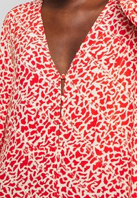 Červené a krémové šaty s potiskem, které mají hluboký V-výstřih, zapínání na knoflíky a abstraktní tvary z lehké tkaniny.