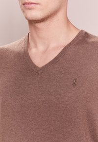 Pull marron à col en V en tissu doux. Présente une texture subtile et un logo brodé sur le côté gauche de la poitrine.