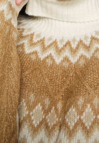 Stickad tröja med en krämfärgad hög hals och en honungsfärgad kropp som har ett geometriskt diamantmönster i olika nyanser av beige.