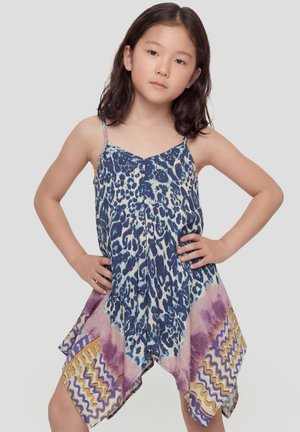 Niña joven de pie con las manos en las caderas, vistiendo un vestido sin mangas con patrones de leopardo azul y abstractos multicolores.