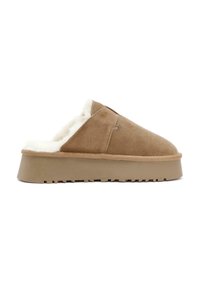 Tan suède slipper met een witte pluizige voering, lage platformzool en een ronde neus. Beschikt over een gladde buitenkant en een gestructureerde zool voor grip.