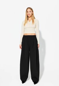 Hellbeige langärmliges Cropped-Top mit zwei Knöpfen am Ausschnitt, kombiniert mit locker sitzenden schwarzen, plissierten Hosen. Glatte Textur.