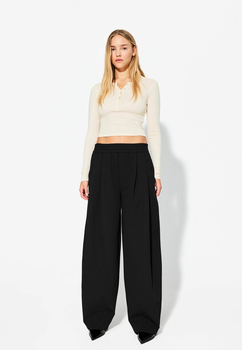 Hellbeige langärmliges Cropped-Top mit zwei Knöpfen am Ausschnitt, kombiniert mit locker sitzenden schwarzen, plissierten Hosen. Glatte Textur.