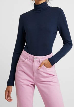 Femme portant un col roulé bleu marine à manches longues, rentré dans un jean taille haute rose clair, une main dans une poche.