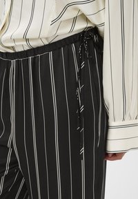 Hlače s črno-belim črtastim vzorcem in elastičnim pasom ter vrvico, v kombinaciji z ujemajočo se črtasto bluzo z gumbnimi manšetami.