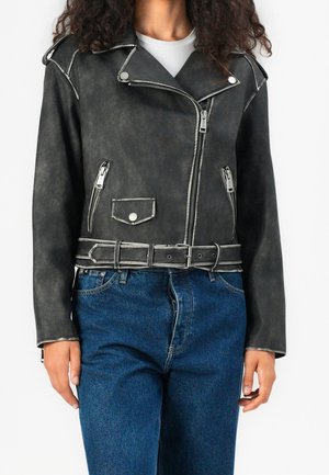 Mujer que lleva una chaqueta de cuero negra de motorista con cremalleras plateadas y cinturón, combinada con vaqueros azules y una camiseta blanca interior.