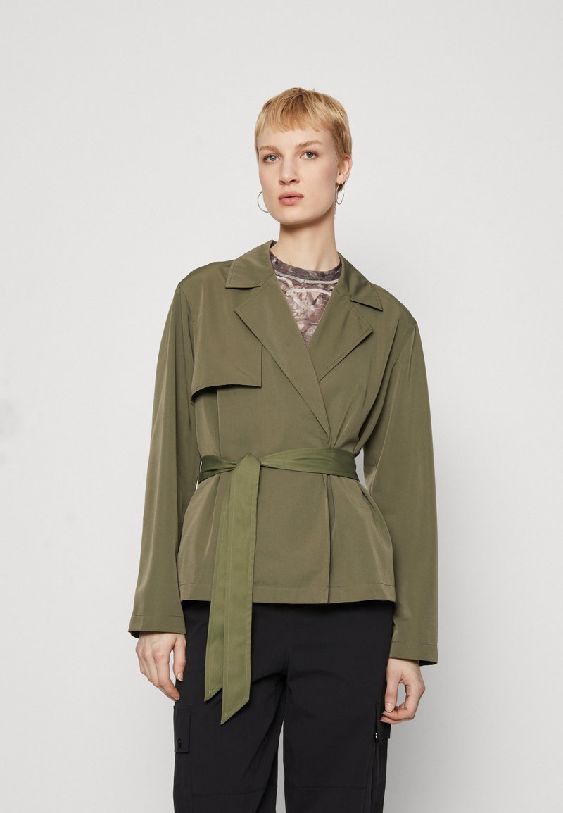 Olive green wrap jacket with a tie belt, featuring a notched collar and front pocket, made of smooth fabric, styled over a patterned top.