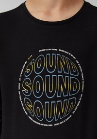 Zwarte t-shirt met korte mouwen met een circulaire graphic met gelaagde tekst in blauw en geel, met duidelijk de tekst "SOUND."