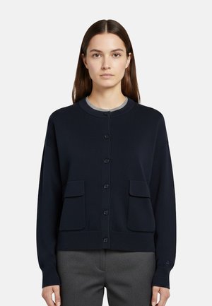 Donna che indossa un cardigan blu navy con bottoni e due tasche frontali sopra una maglietta grigia, in piedi davanti a uno sfondo chiaro e semplice.