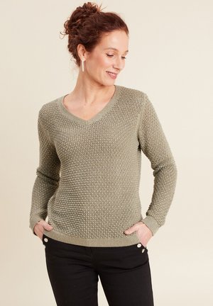 V-Ausschnitt Strickpullover in hellolivgrün mit strukturiertem Design, gerippten Bündchen und Saum. Kombiniert mit schwarzen Hosen mit silbernen Knöpfen.