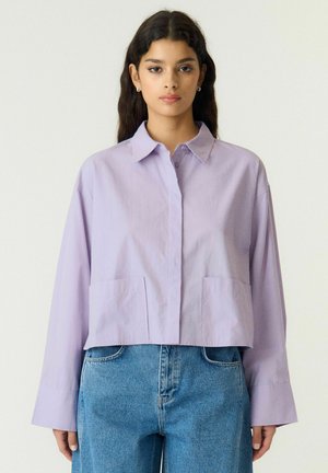 Jeune femme aux longs cheveux foncés portant une chemise lilas clair à col et un jean bleu taille haute, debout devant un fond clair uni.