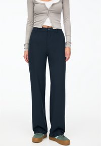 Pantalons navy à jambes larges en tissu lisse, avec une fermeture à bouton, des poches avant et une coupe ajustée, associés à des baskets vertes.