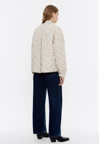 Giacca bomber trapuntata beige con colletto alto e polsini a costine, indossata sopra jeans larghi scuri e stivali neri con tacco.
