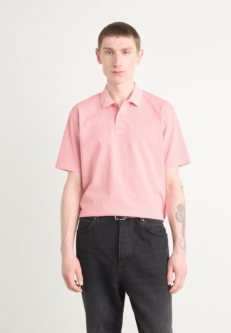Polo majica s kratkimi rokavi v rožnati barvi s kragnico; ima belo vezeno logotipo na prsih. Dopolnjena s temnimi jeans hlačami.