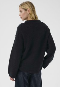 Černý pletený svetr s texturou a volným střihem, kulatým výstřihem a spadlými rameny. Design zahrnuje dlouhé rukávy a mírně oversized siluetu.