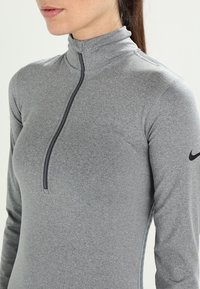 Harmaa, pitkähihaisella urheilupaidalla, jossa on neljännesvetoketju, korkea kaulus ja Nike-logo vasemmassa hihassa. Sileä, joustava kangas.