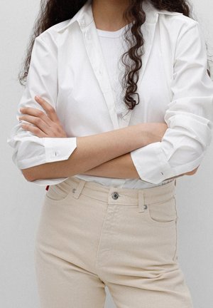 Henkilö, jolla on pitkät kiharat hiukset, pukeutunut valkoiseen nappikauluspaitaan valkoisen topin päällä, beigeihin korkeavyötäröisiin housuihin, kädet ristissä.