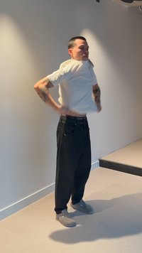 Mann mit kurzen Haaren zieht in einem minimalistisch eingerichteten Raum mit hellen Wänden, schwarzer Hose und grauen Schuhen ein weißes T-Shirt über den Kopf.