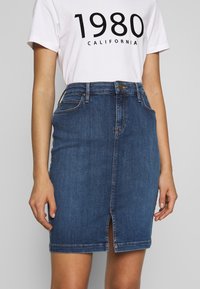 Jeanpennkjol i mörkblått med en framklips, fem-fickorsdesign och kontrastsöm. Bärs med en vit t-shirt med tryck.